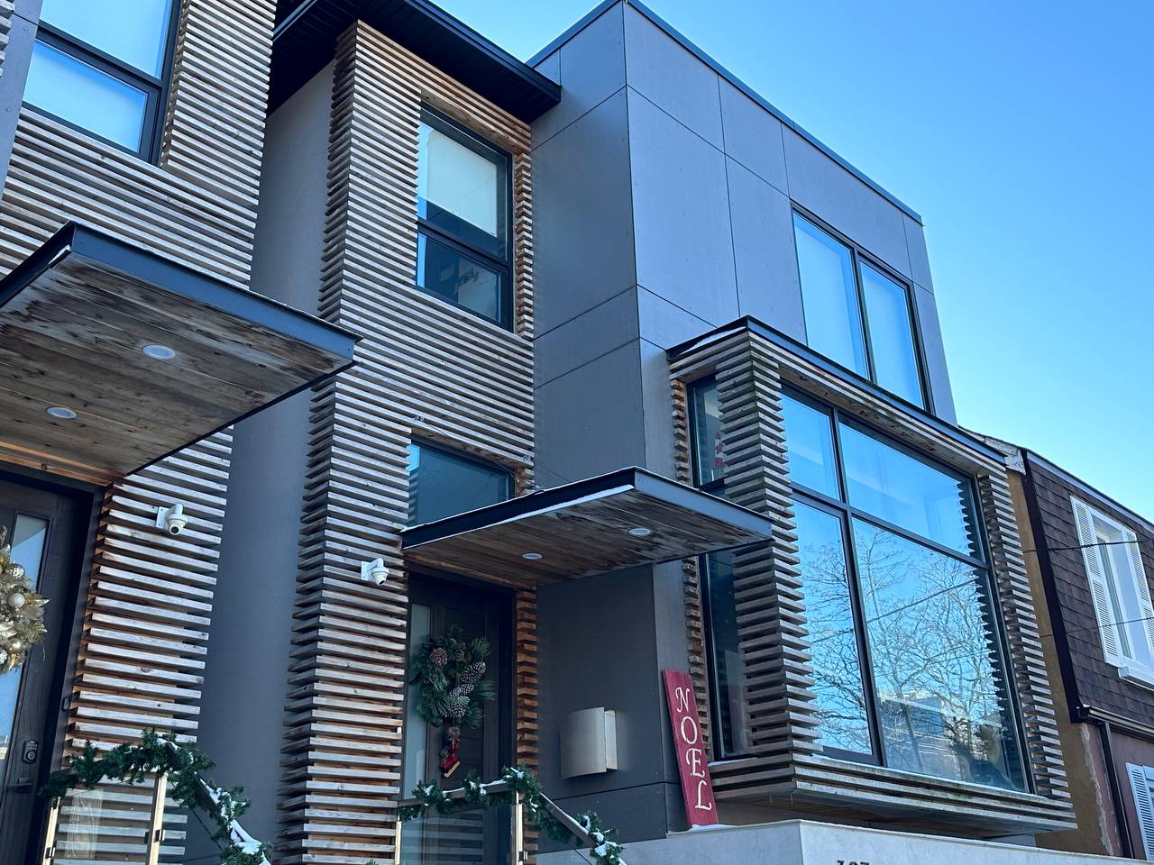 Modern house exterior with wooden slat detailing, large windows, and a holiday wreath, under a clear blue sky.