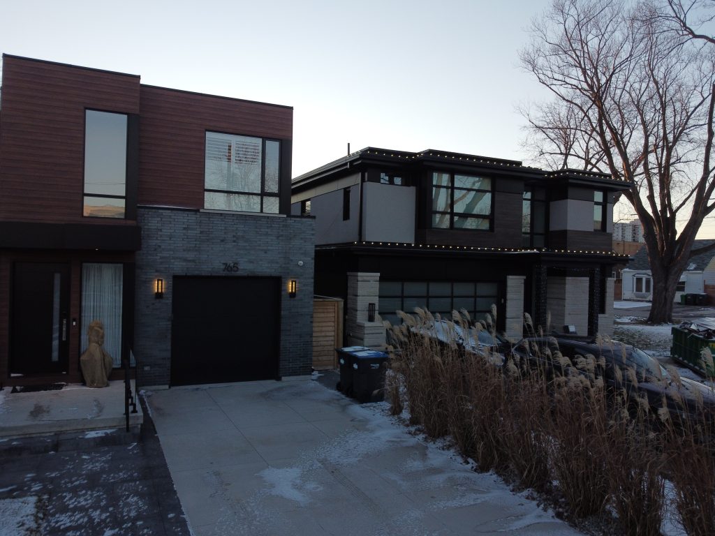 Two contemporary, cubic-style homes at dusk with cars parked in the driveways. The house on the left combines grey brick on the ground level with dark brown horizontal wood-look aluminum siding on the upper box.