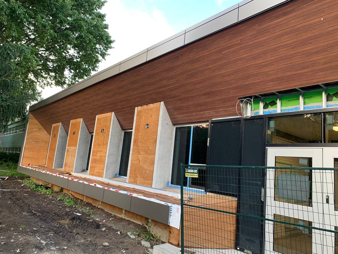 Rear view construction site of a long institutional building. The upper facade features dark walnut horizontal wood-look aluminum siding above grey panels. A vehicle is partially visible in the distance on the far left.